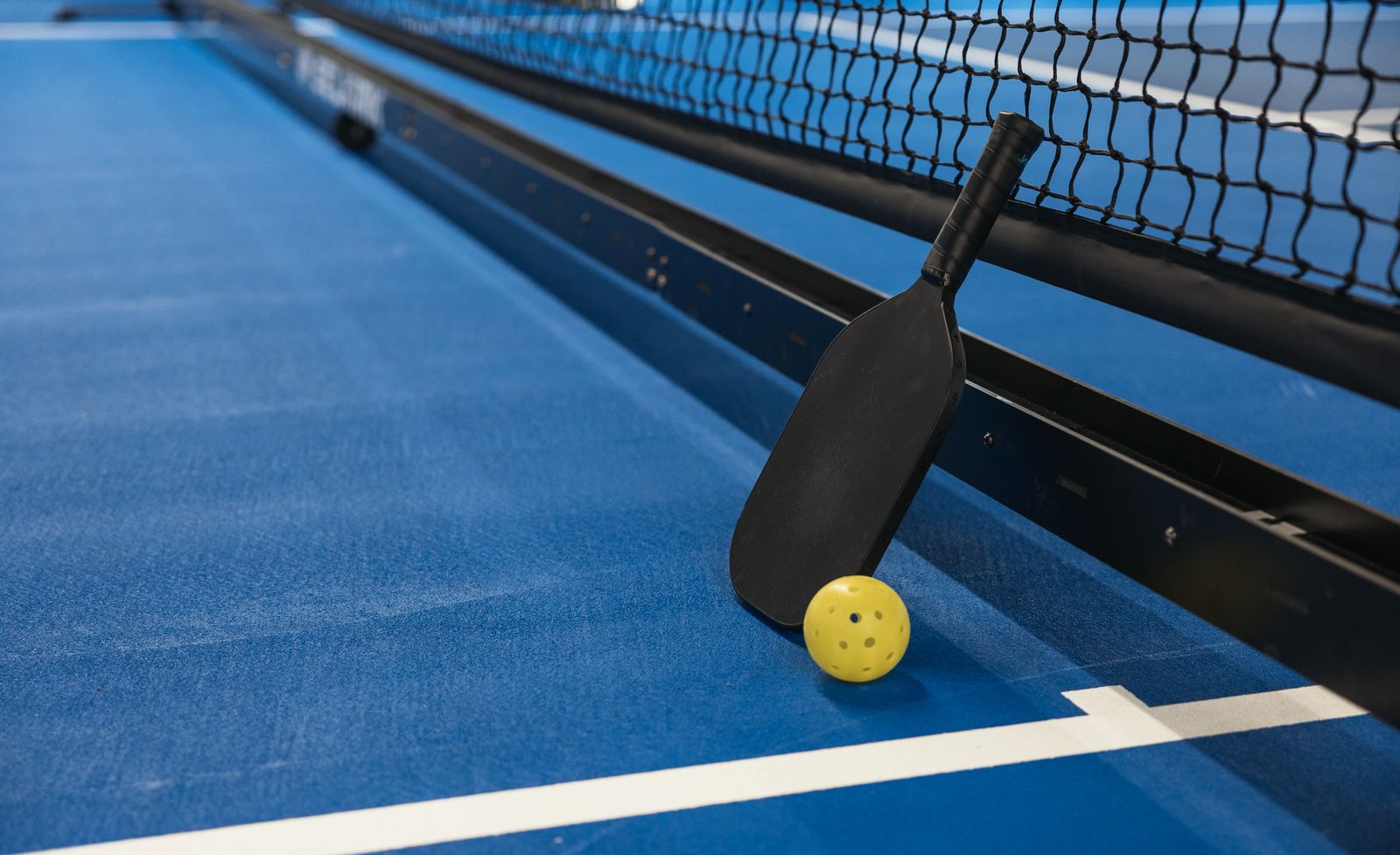 A pickleball paddle and ball resting against the net on a blue court